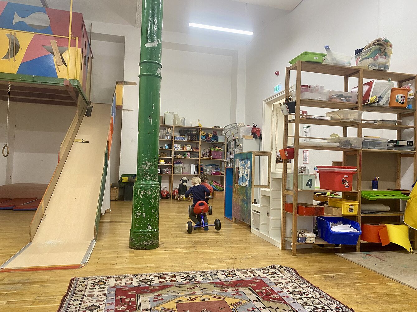 Kind auf Dreirad in einem hellen Spielzimmer mit Rutsche, Regalen und buntem Teppich