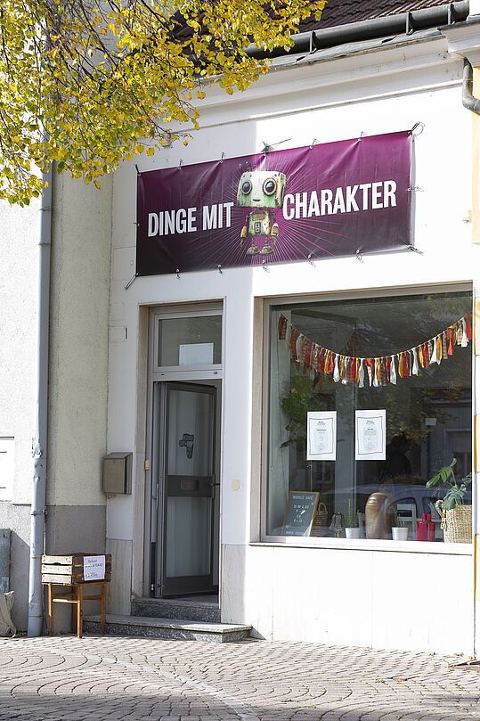Foto: Susanne Senekowitsch Ladenfront mit offenem Eingang, darüber ein violettes Banner mit der Aufschrift 'DINGE MIT CHARAKTER' und einer bunten Roboterfigur.