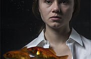 Eine weiblich zu lesende Person steht hinter einem Aquarium in dem Goldfische schwimmen.