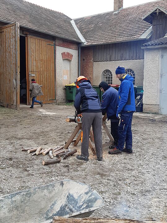 Drei Personen stehen um eine Sägebock mit einer Kettensäge und schneiden Holzstücke in einem Hof mit alten Gebäuden im Hintergrund