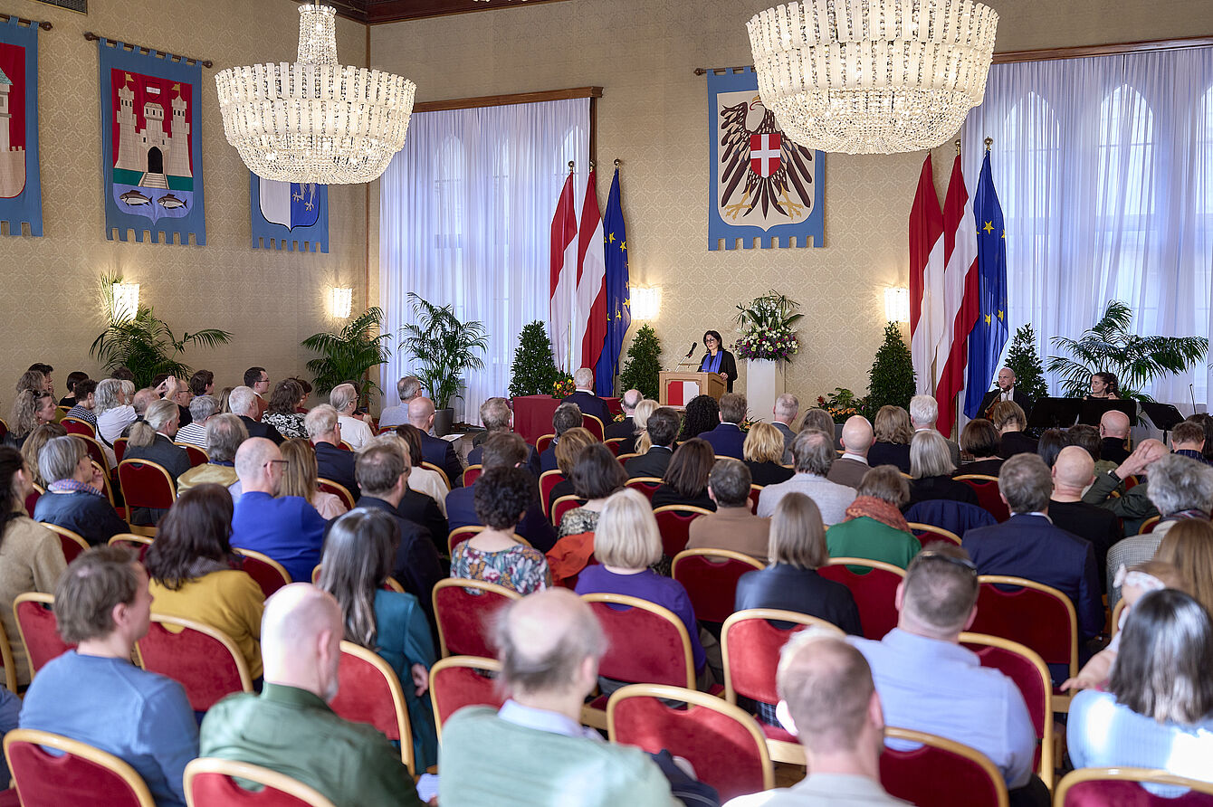 Großer Saal mit Publikum auf roten Stühlen vor einer Rednerin am Podium, umgeben von Fahnen und Kronleuchtern