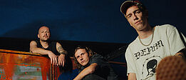 Die Band Anda Morts, drei männlich gelesene Personen, blicken sitzend Richtung Kamera, dahinter Abendhimmel und eine verrostete Fläche zwischen ihnen.