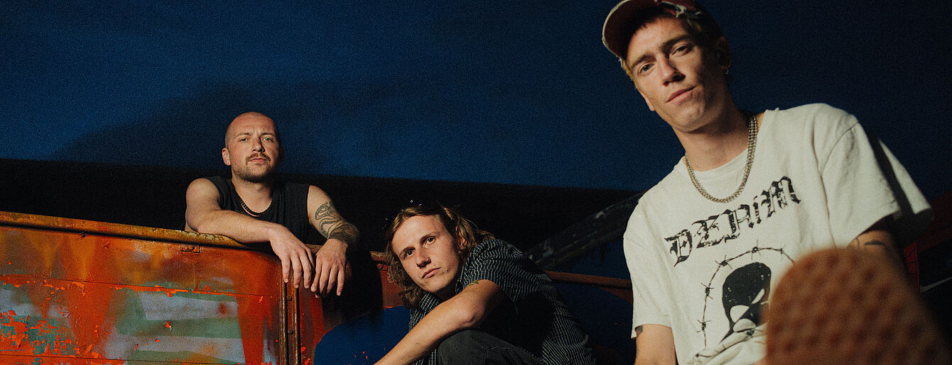 Die Band Anda Morts, drei männlich gelesene Personen, blicken sitzend Richtung Kamera, dahinter Abendhimmel und eine verrostete Fläche zwischen ihnen.
