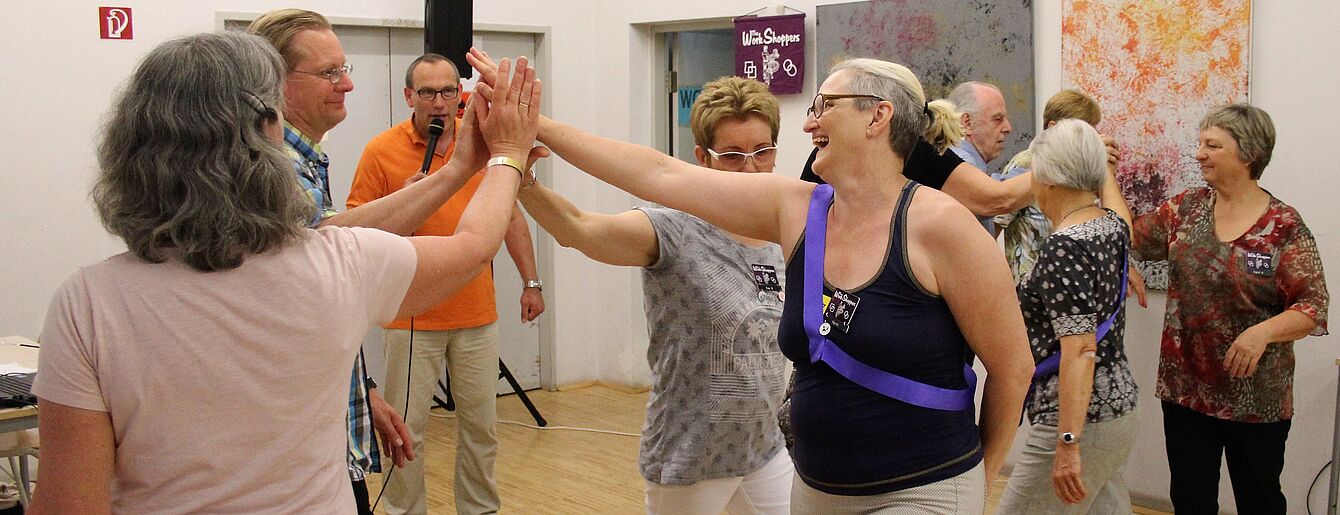 Gruppe älterer Menschen tanzt in einem hellen Raum mit Holzfußboden und abstrakten Bildern an den Wänden