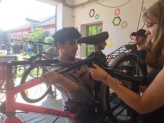 Eine Person hält ein Fahrrad, während eine Person ihm hilft, das Fahrrad zu reparieren. Im Hintergrund sind weitere Fahrräder und Menschen sichtbar.