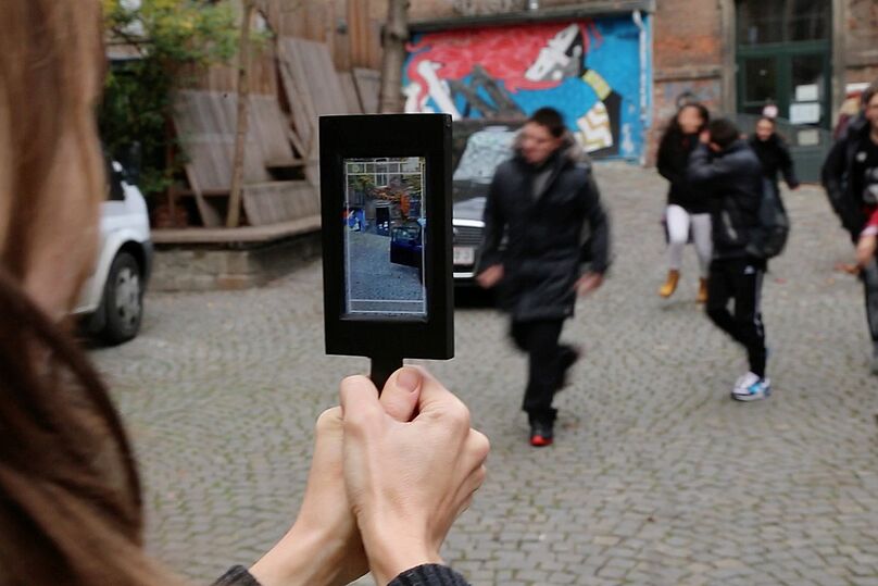 Person hält Tablet mit Kameraansicht auf eine Gruppe von Menschen auf einem gepflasterten Platz