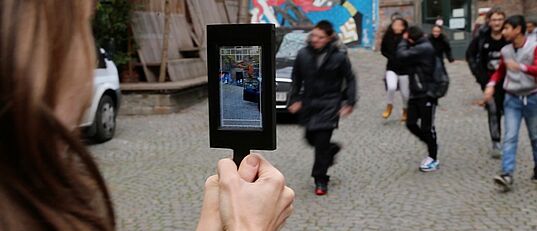 Person hält Tablet mit Kameraansicht auf eine Gruppe von Menschen auf einem gepflasterten Platz