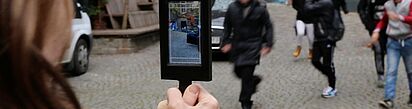 Person hält Tablet mit Kameraansicht auf eine Gruppe von Menschen auf einem gepflasterten Platz
