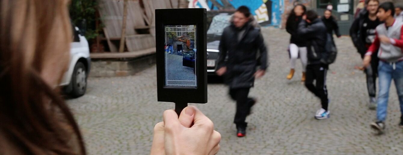 Person hält Tablet mit sichtbarem Bild von sich selbst, im Hintergrund mehrere Personen auf gepflastertem Platz