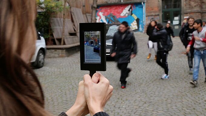 Person hält Tablet mit Kameraansicht auf eine Gruppe von Menschen auf einem gepflasterten Platz