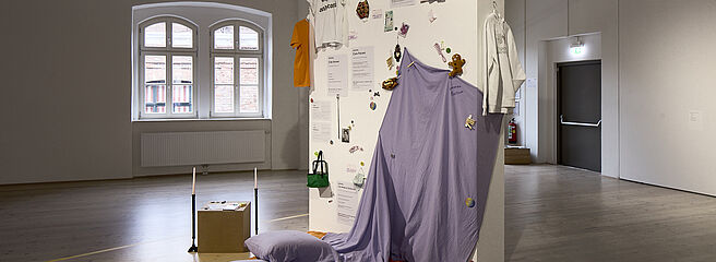 Das Bild zeigt eine Ausstellungsobjekt mit einem lila Tuch an einer weißen Wand, im Hintergrund zwei Fenster