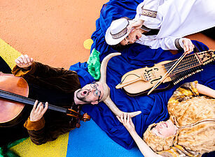 Drei Personen liegen auf buntem Boden, eine spielt Cello, eine hält ein historisches Streichinstrument, die dritte trägt ein Fellkleid und hält ein Horn.