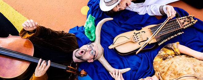 Drei Personen liegen auf buntem Boden, eine spielt Cello, eine hält ein historisches Streichinstrument, die dritte trägt ein Fellkleid und hält ein Horn.