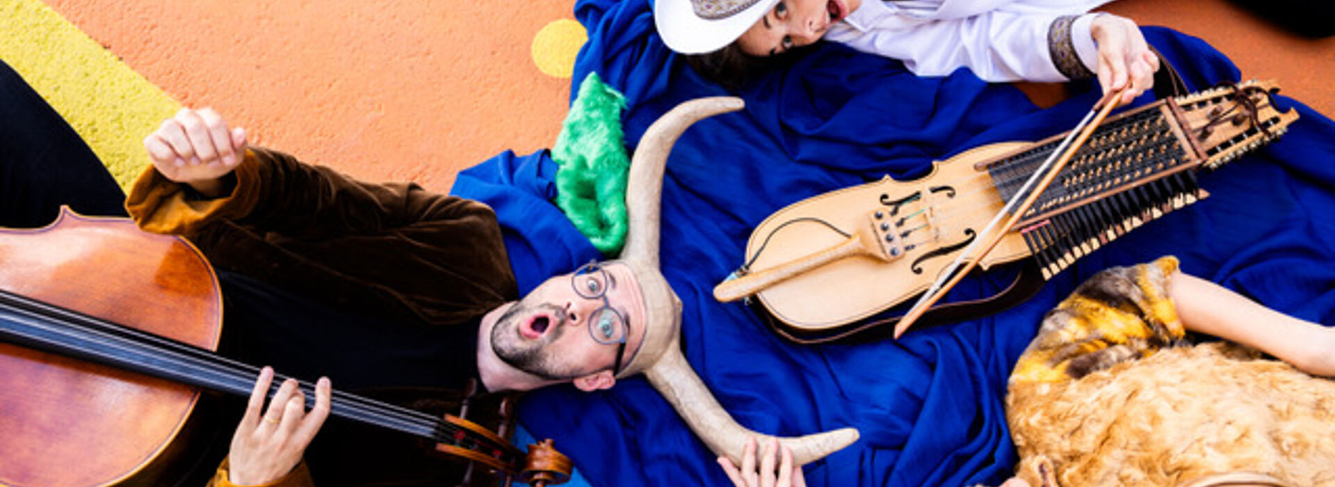 Drei Personen liegen auf buntem Boden, eine spielt Cello, eine hält ein historisches Streichinstrument, die dritte trägt ein Fellkleid und hält ein Horn.