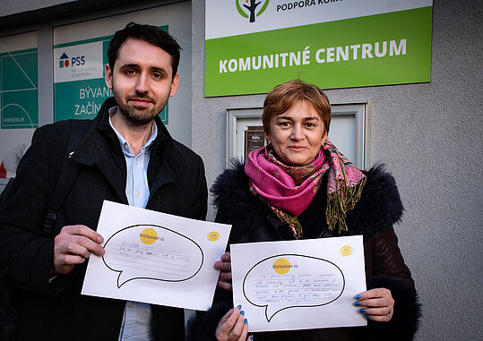 Zwei Menschen mit Papieren mit der Aufschrift "Inclusion is" vor dem Komunitné Centrum in Bratislava