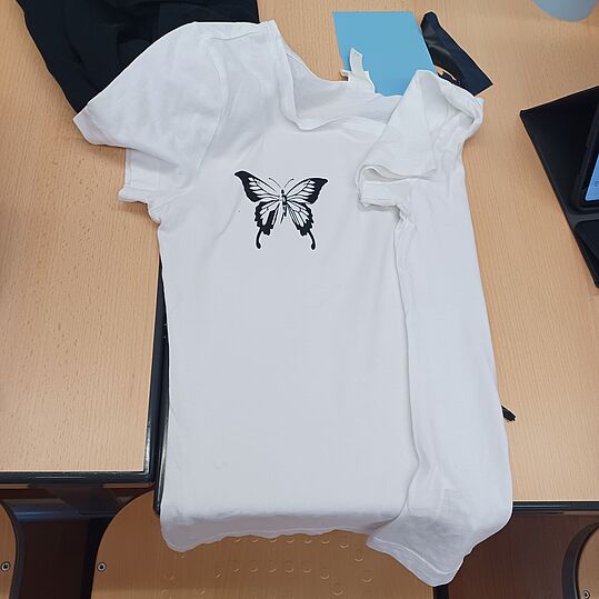 Weiße T-Shirt mit schwarzem Schmetterlingsmotiv liegt auf einem hellen Tisch neben einem weißen Kleidungsstück und einem Laptop