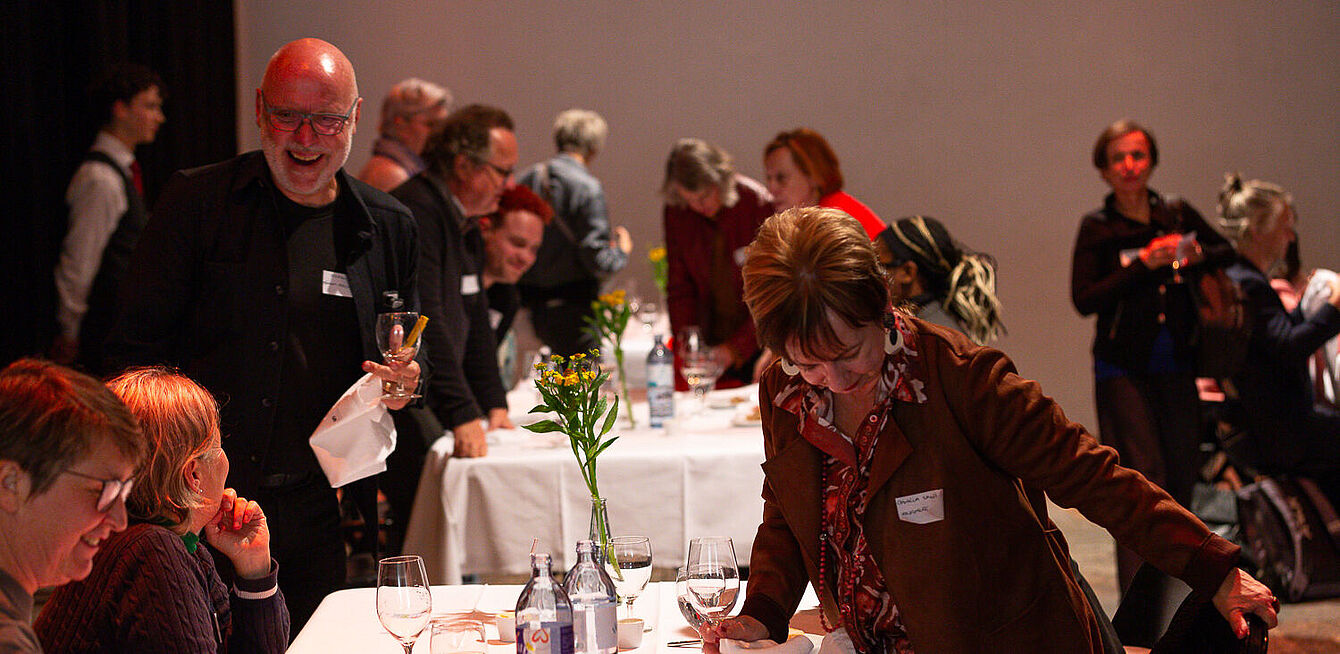 Foto: Susanne Senekowitsch Mehrere Personen stehen und sitzen an einem gedeckten Tisch mit weißen Tischdecken, Gläsern, Wasserflaschen und einer Blumenvase in einem Veranstaltungsraum.