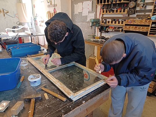 Zwei Personen arbeiten an einem alten Fensterrahmen auf einer Werkbank in einer Werkstatt