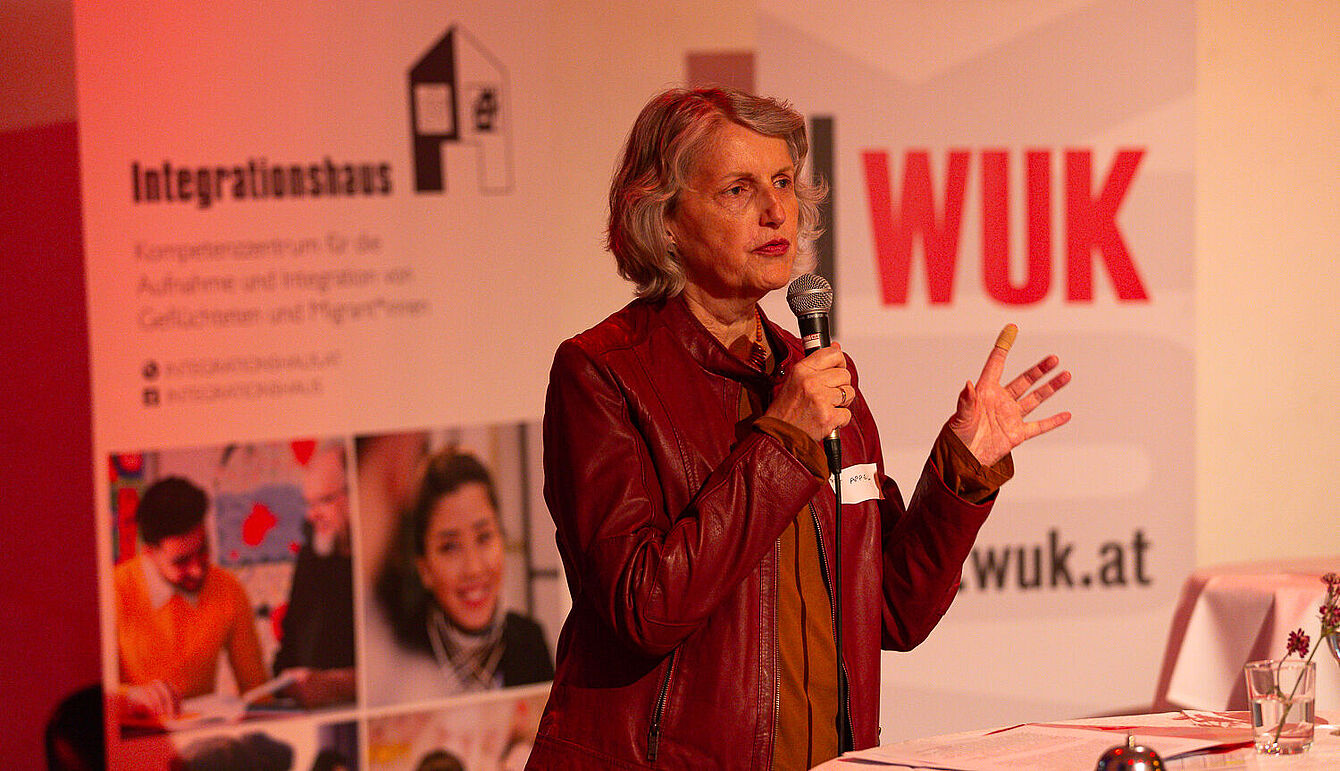 Foto: Susanne Senekowitsch Eine Frau mit roter Lederjacke hält Mikrofon und spricht vor Bannern von Integrationshaus und WUK.