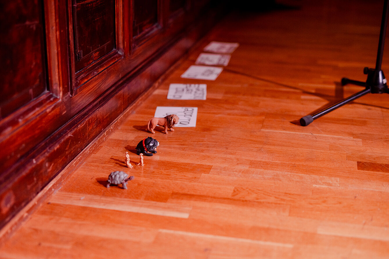 Mehrere kleine Tierfiguren und Zettel liegen in einer Reihe auf einem Holzboden neben einer Holzvertäfelung.