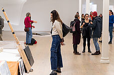 Mehrere Personen in einer Galerie betrachten Kunstwerke und unterhalten sich, eine Frau steht nah an einer Wand mit aufgestellten Bildern und Objekten