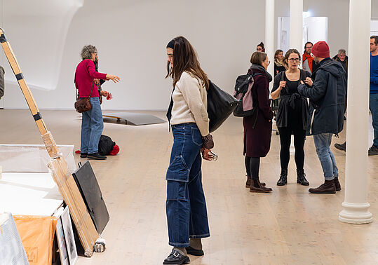Mehrere Personen in einer Galerie betrachten Kunstwerke und unterhalten sich, eine Frau steht nah an einer Wand mit aufgestellten Bildern und Objekten