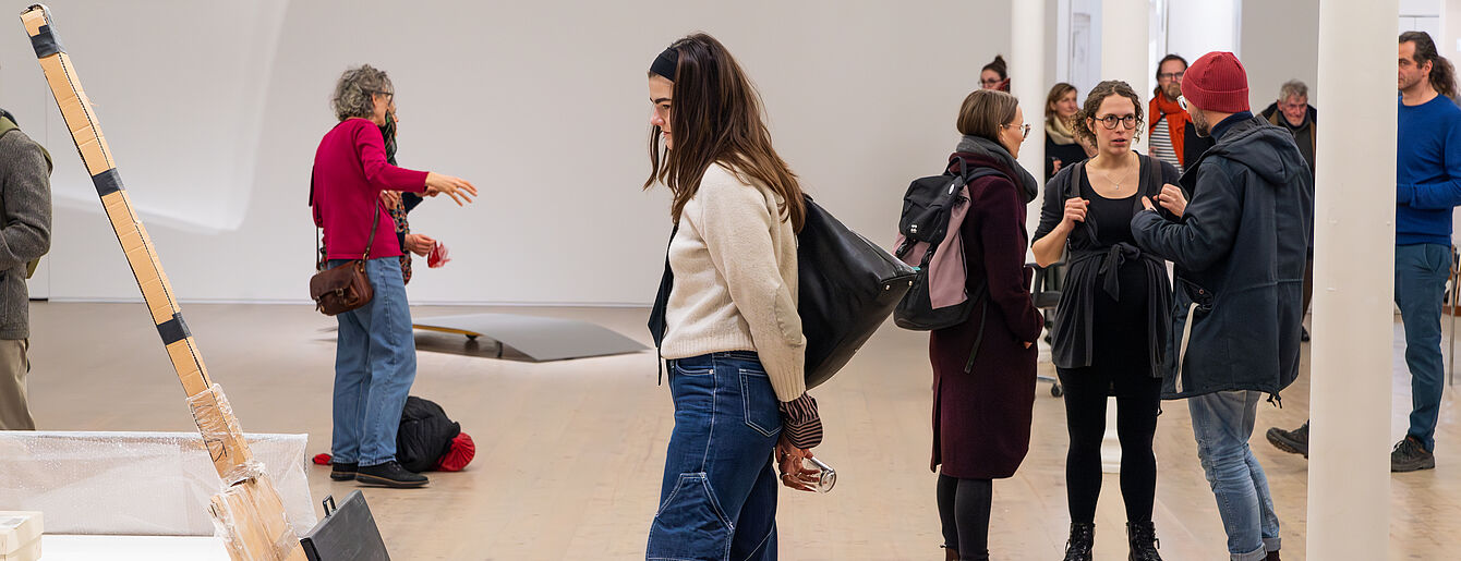 Mehrere Personen in einer Galerie betrachten Kunstwerke und unterhalten sich, eine Frau steht nah an einer Wand mit aufgestellten Bildern und Objekten