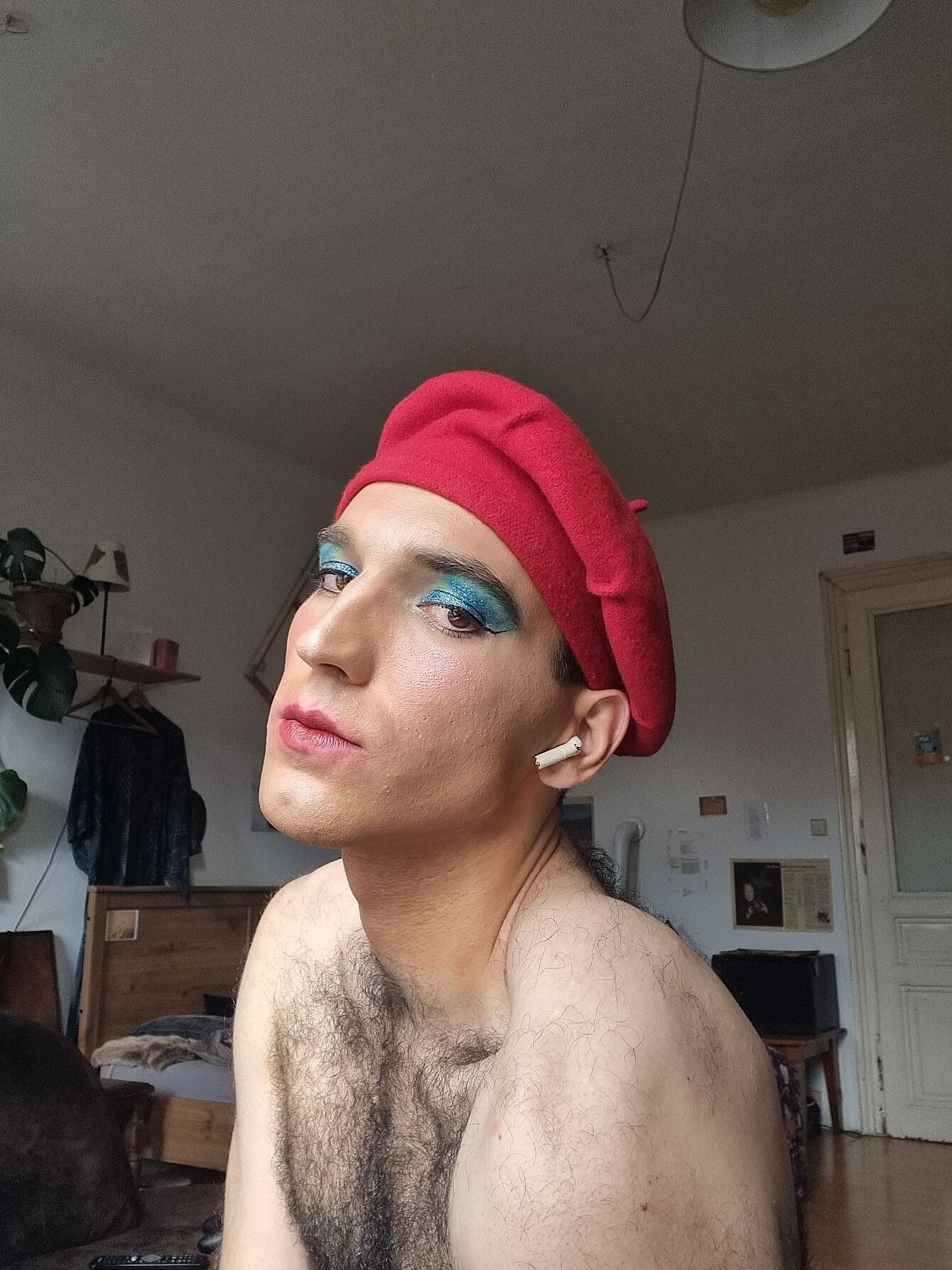Person mit roter Baskenmütze, blauem Lidschatten und nackten Schultern in einem Zimmer mit Möbeln im Hintergrund