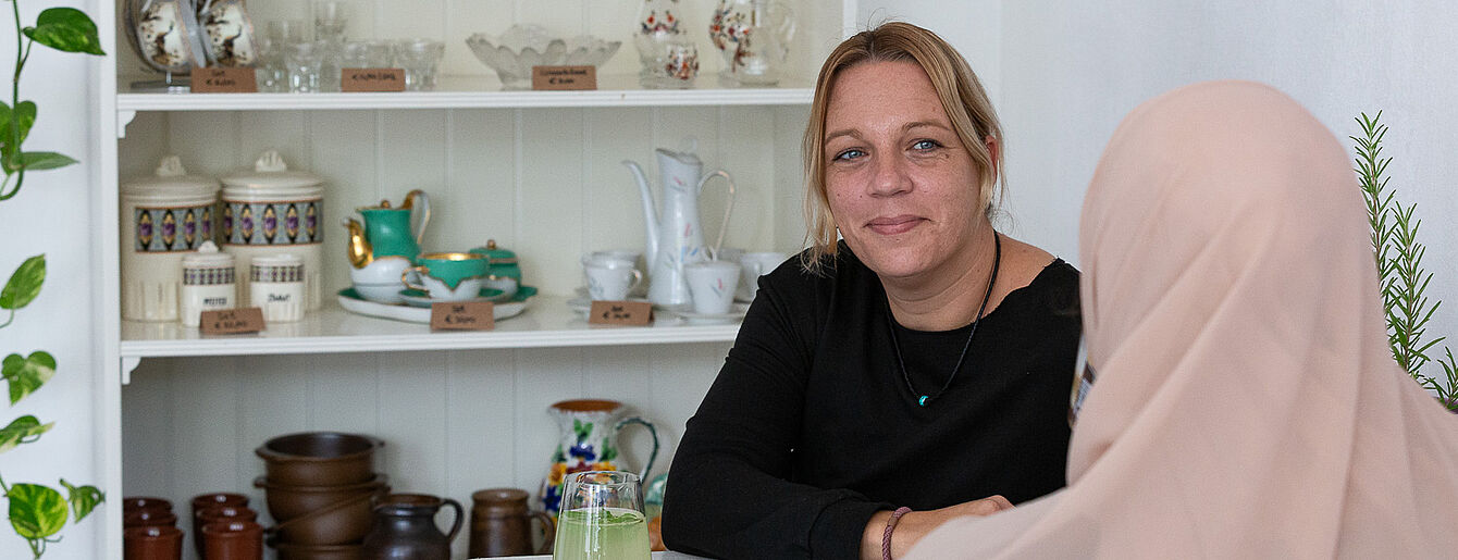 Foto: Susanne Senekowitsch Zwei Frauen sitzen an einem weißen Tisch, im Hintergrund ein Regal mit Keramikgeschirr und Gläsern