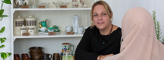 Foto: Susanne Senekowitsch Zwei Frauen sitzen an einem weißen Tisch, im Hintergrund ein Regal mit Keramikgeschirr und Gläsern