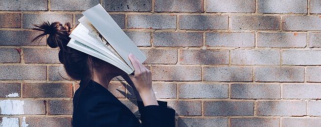 Eine Frau steht mit Büchern vor dem Gesicht vor einer Ziegelwand