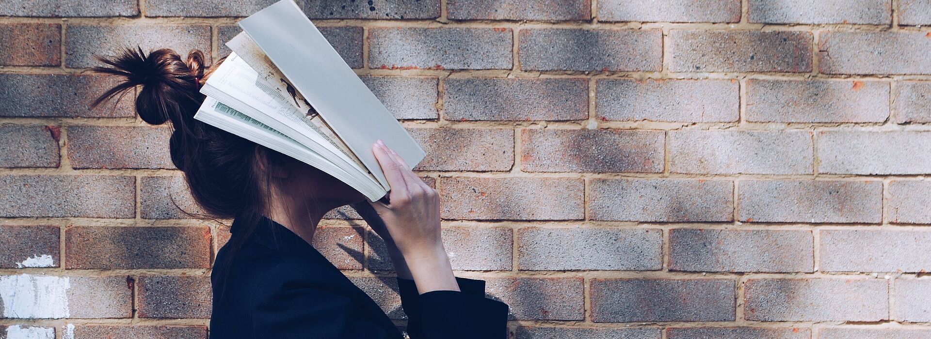 Eine Frau steht mit Büchern vor dem Gesicht vor einer Ziegelwand