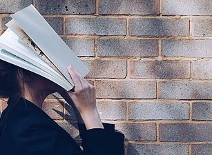Eine Frau steht mit Büchern vor dem Gesicht vor einer Ziegelwand