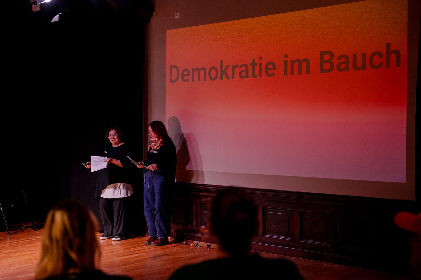 Zwei Personen stehen auf einer Bühne vor einer Wand mit der Projektion 'Demokratie im Bauch', im Vordergrund sind Zuschauer*innen unscharf zu sehen.