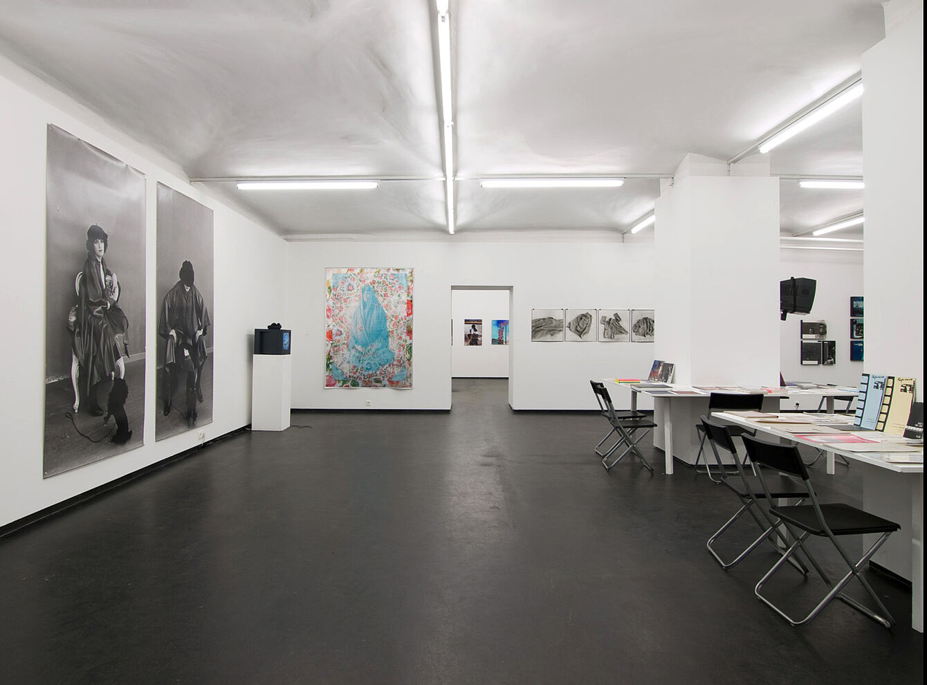 Weißer Ausstellungsraum mit großen Schwarz-weiß-Porträts an der linken Wand, farbigem Bild in der Mitte und mehreren Zeichnungen an der rechten Wand, Tische mit Stühlen rechts