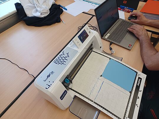 Schneideplotter auf einem Tisch mit einem Laptop daneben und einer Person, die darauf tippt
