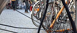 Nahaufnahme eines Fahrradhinterrads mit Speichen und Kettenritzel vor einem gepflasterten Platz mit mehreren Fahrrädern und Personen im Hintergrund