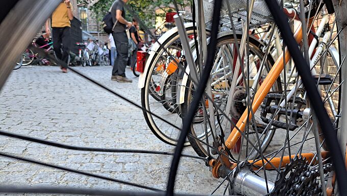Nahaufnahme eines Fahrradhinterrads mit Speichen und Kettenritzel vor einem gepflasterten Platz mit mehreren Fahrrädern und Personen im Hintergrund