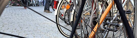 Nahaufnahme eines Fahrradhinterrads mit Speichen und Kettenritzel vor einem gepflasterten Platz mit mehreren Fahrrädern und Personen im Hintergrund
