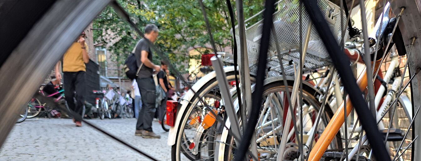 Nahaufnahme eines Fahrradhinterrads mit Speichen und Kettenritzel vor einem gepflasterten Platz mit mehreren Fahrrädern und Personen im Hintergrund