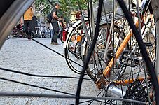 Nahaufnahme eines Fahrradhinterrads mit Speichen und Kettenritzel vor einem gepflasterten Platz mit mehreren Fahrrädern und Personen im Hintergrund