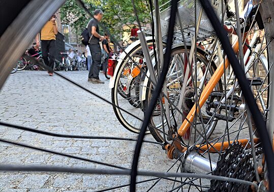 Nahaufnahme eines Fahrradhinterrads mit Speichen und Kettenritzel vor einem gepflasterten Platz mit mehreren Fahrrädern und Personen im Hintergrund