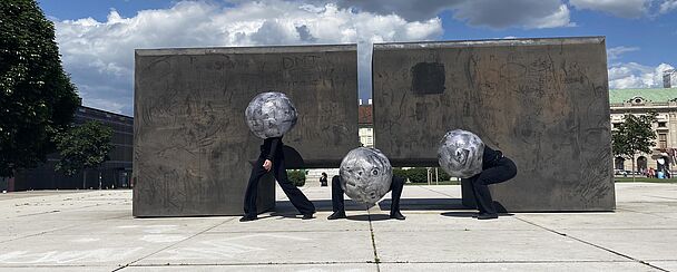 Drei Personen mit kugelförmigen Helmen in einer urbanen Umgebung vor zwei großen rechteckigen Betonwänden unter bewölktem Himmel