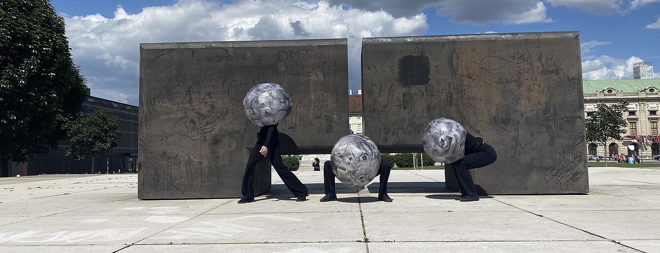 Drei Personen mit kugelförmigen Helmen in einer urbanen Umgebung vor zwei großen rechteckigen Betonwänden unter bewölktem Himmel