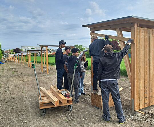 Sechs Personen arbeiten an Holzstrukturen im Freien. Im Hintergrund sind weitere Holzrahmen aufgestellt. Der Boden ist unbefestigt.