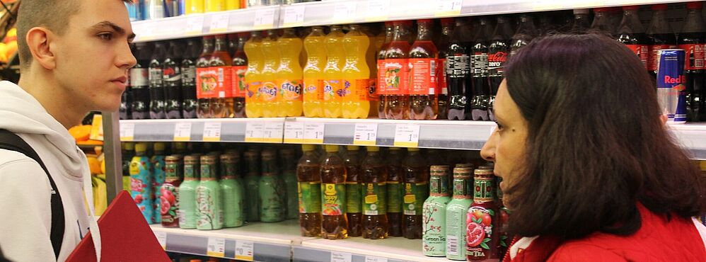 Jugendliche Person und andere Person im Gespräch vor einem Regal mit verschiedenen Getränkeflaschen im Supermarkt