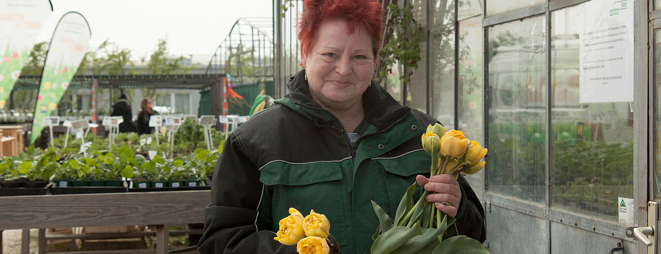 Person mit roten Haaren hält gelbe Tulpen in einem Gewächshaus mit Pflanzen im Hintergrund