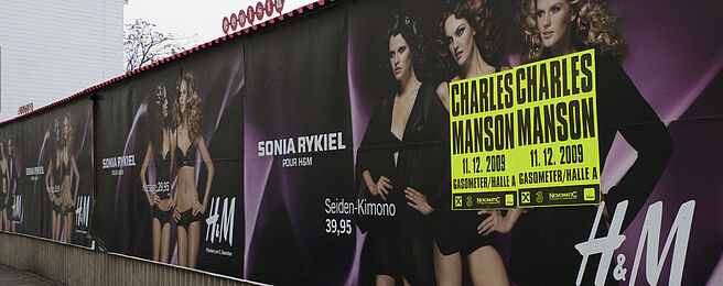 Foto von einer Gewista-Plakatwand, auf die ein neongelbes Konzertplakat von Charles Manson angebracht wurde. 