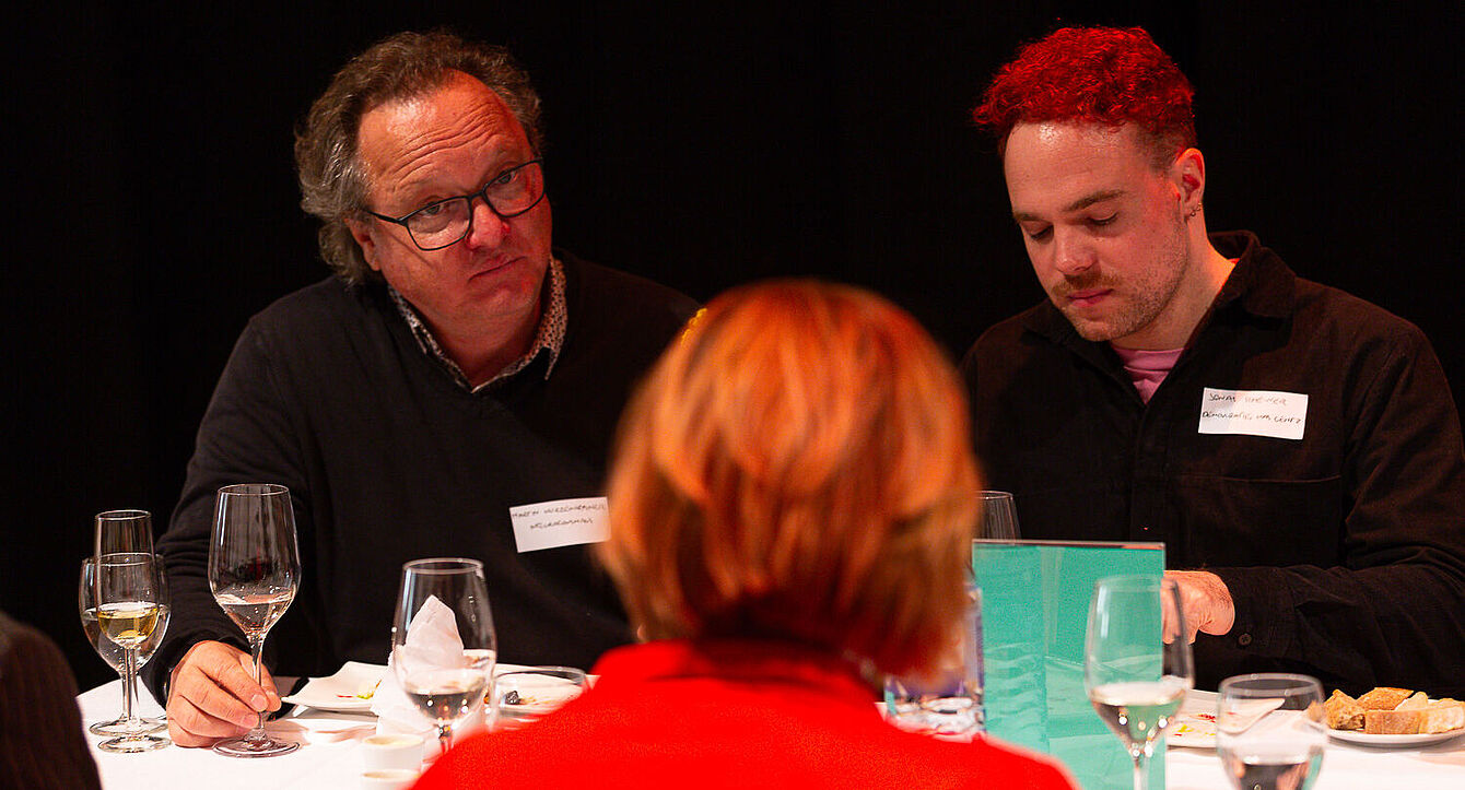 Foto: Susanne Senekowitsch Drei Personen sitzen an einem Tisch mit Gläsern, Tellern und einer grünen Tafel, eine Person trägt roten Pullover.
