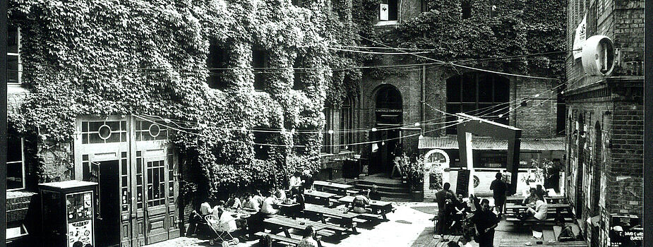 Innenhof mit mehreren Holzbänken und Tischen, an denen Menschen sitzen und gehen, umgeben von Gebäuden mit von wildem Wein bewachsenen Wänden